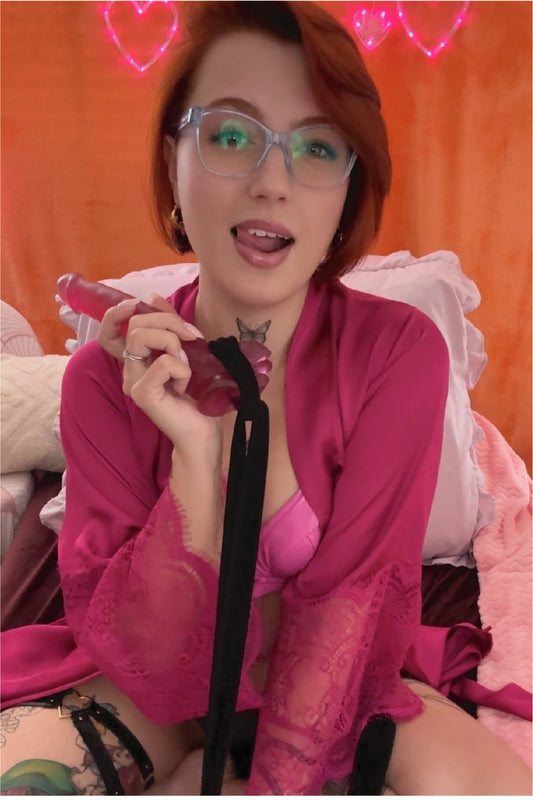 Dame in roze lingerie en kimono houdt een roze seksspeeltje vast in een slaapkamer met hartvormige verlichting